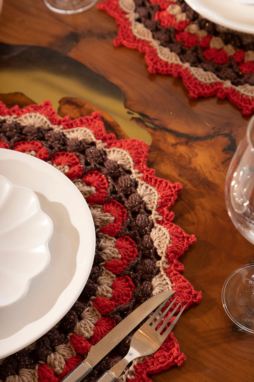 Círculo Barroco Maxcolor Sparkle – Cotton Yarn (216m / 200g) - A close-up of a handmade crocheted placemat with a scalloped edge in red, brown, and beige, styled on a rustic wooden table with white plates and a fork.