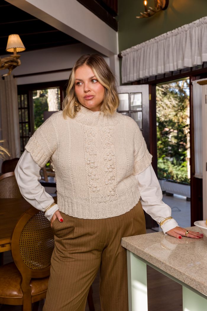 Circulo InLove - 100% Brazilian Cotton Yarn (125m/100g)- A woman wearing a cream cable-knit mock neck sweater vest over a white long-sleeve shirt and brown pants, standing in a home interior near a kitchen island.