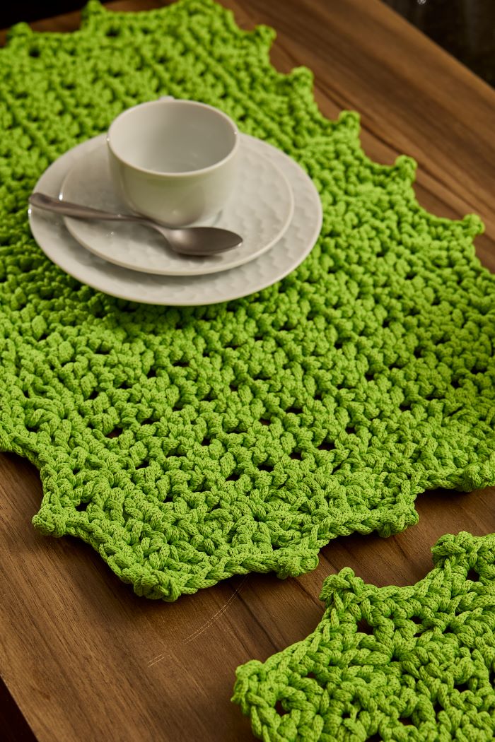 Circulo Fio Náutico Poly 5 mm – Polyester Cord Yarn ( 178 m / 500 g) - A bright lime green crocheted placemat with an irregular scalloped edge, styled on a wooden table with a white ceramic cup and saucer and a spoon.