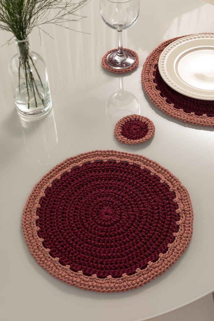 Circulo Fio Náutico Poly 5 mm – Polyester Cord Yarn ( 178 m / 500 g) - A set of hand-crocheted round placemats and coasters in burgundy and dusty rose, styled on a white table with plates, a wine glass, and a vase of greenery.